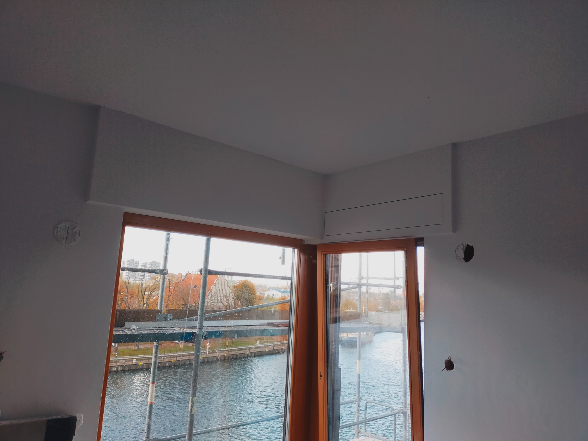 Wnętrze w trakcie remontu z widokiem na rzekę przez okno, widoczne ślady po instalacjach na ścianie i zabudowa sufitowa.