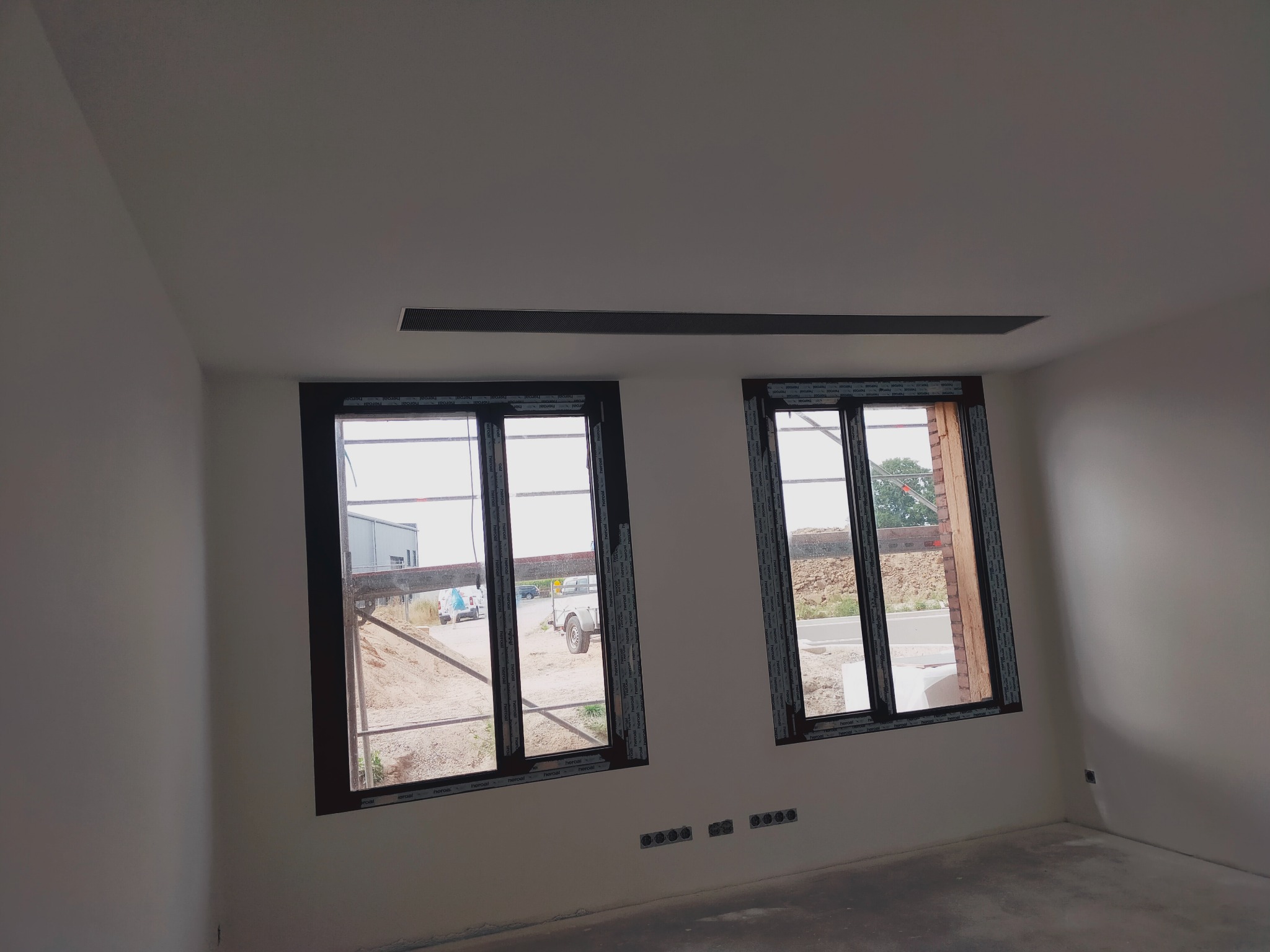 Wnętrze w trakcie wykończenia z dwoma oknami z brązowymi ramami, widoczne nieotynkowane ściany zewnętrzne i przygotowane otwory na gniazdka elektryczne.