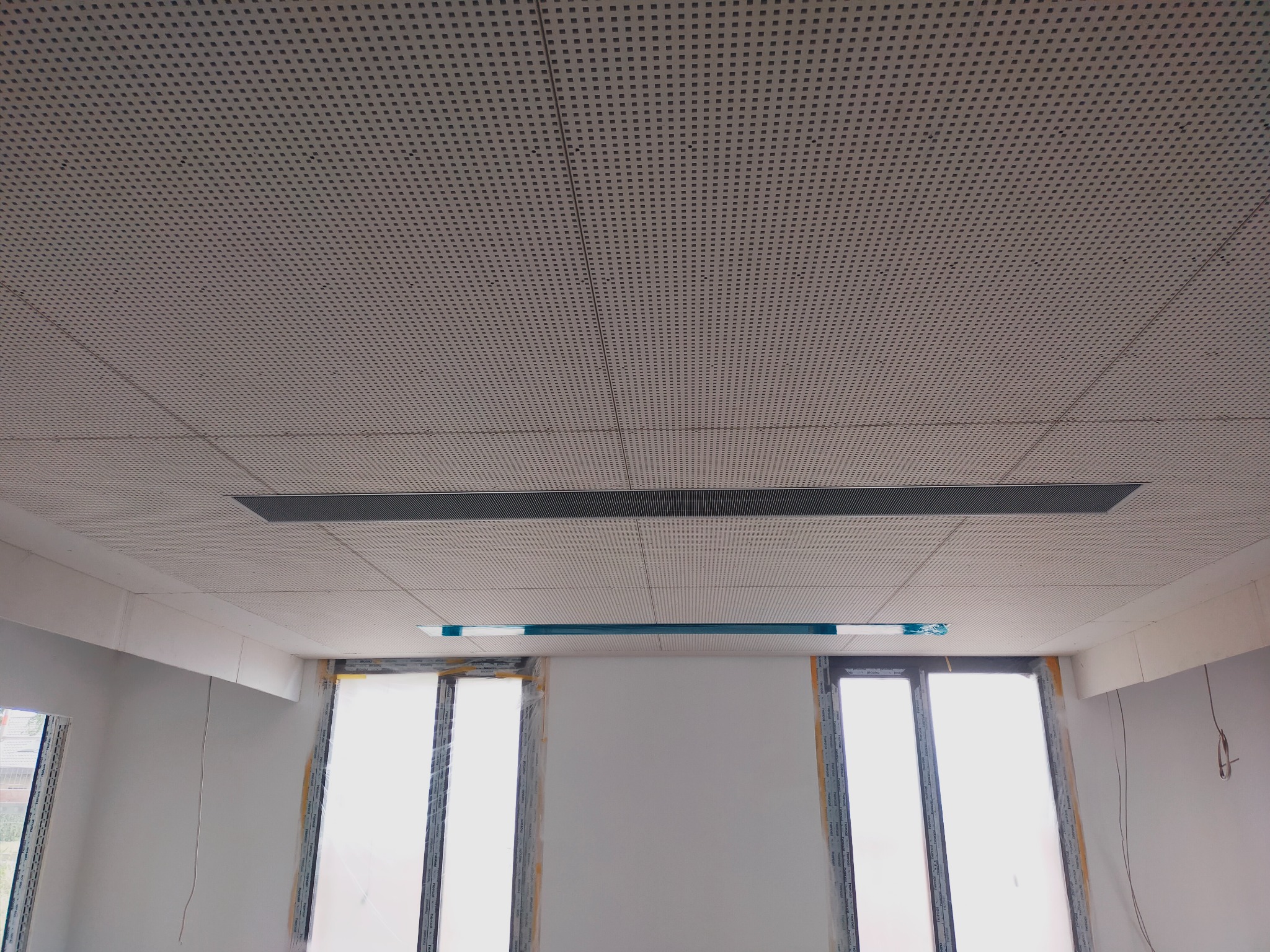 Sufit podwieszany z perforowanej płyty w pomieszczeniu w trakcie wykańczania, widoczne okna zabezpieczone folią.