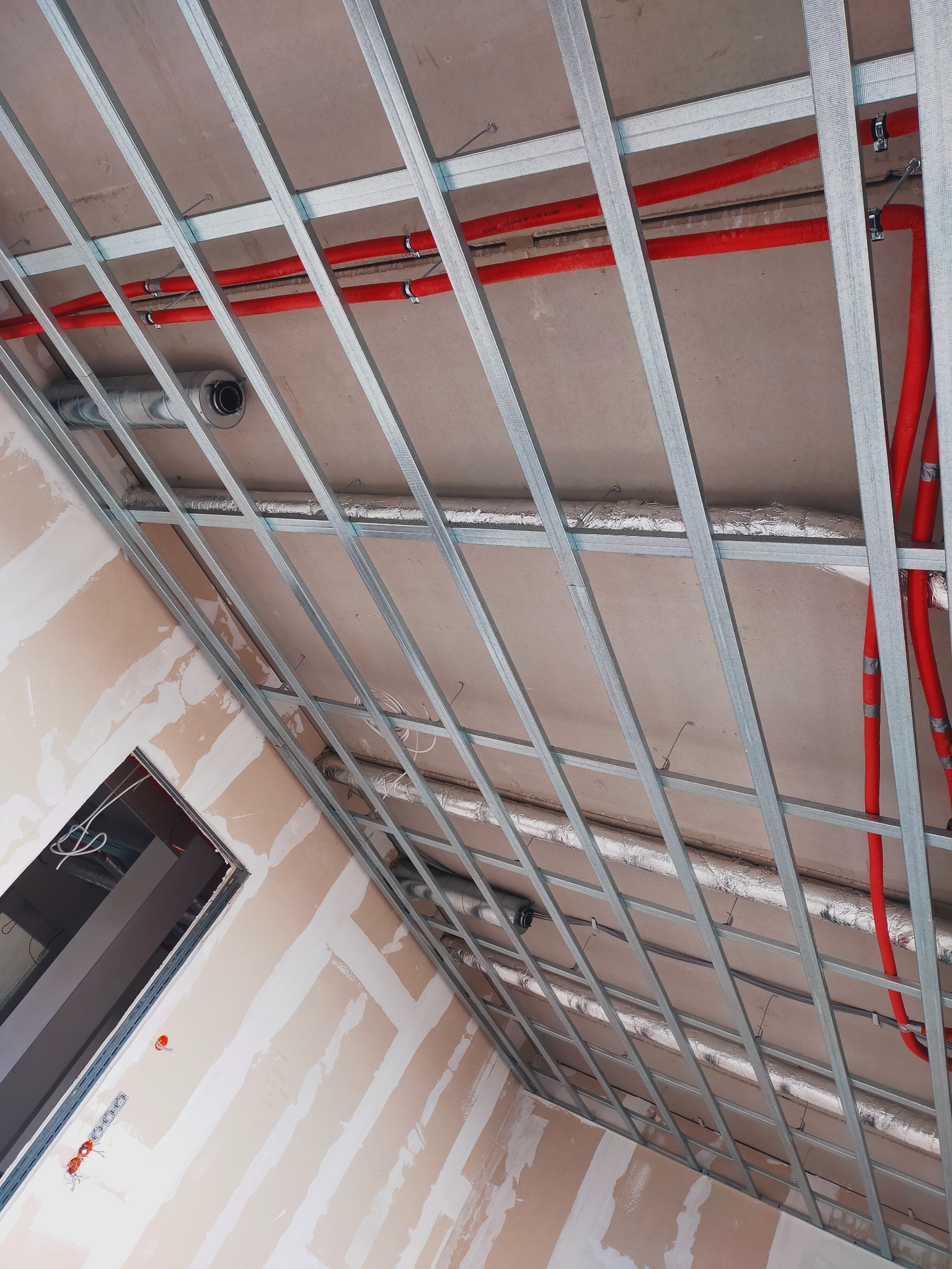 Sufit podwieszany ze stalową konstrukcją nośną, widoczne czerwone rury instalacyjne i przewody wentylacyjne, ściany częściowo wykończone z widocznymi łączeniami płyt.