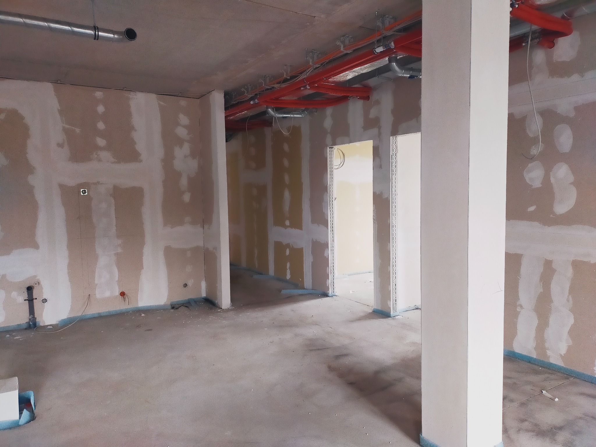 Wnętrze w trakcie prac wykończeniowych: ściany z widocznymi pasami szpachli, instalacje pod sufitem, surowa podłoga zabezpieczona folią przy ścianach, białe filary.