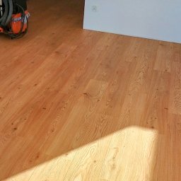 Intra Floor - montaż i naprawa podłóg - Jasny panel MDF ułożony w pokoju, widoczny odkurzacz i narzędzia w tle, cień na podłodze.