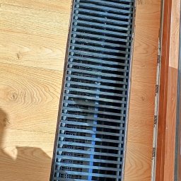 Intra Floor - montaż i naprawa podłóg - Szczegółowe ujęcie liniowego odpływu podłogowego wkomponowanego w jasne panele podłogowe, z widoczną krawędzią okna po prawej stronie.