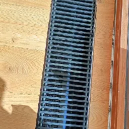 Szczegółowe ujęcie liniowego odpływu podłogowego wkomponowanego w jasne panele podłogowe, z widoczną krawędzią okna po prawej stronie.