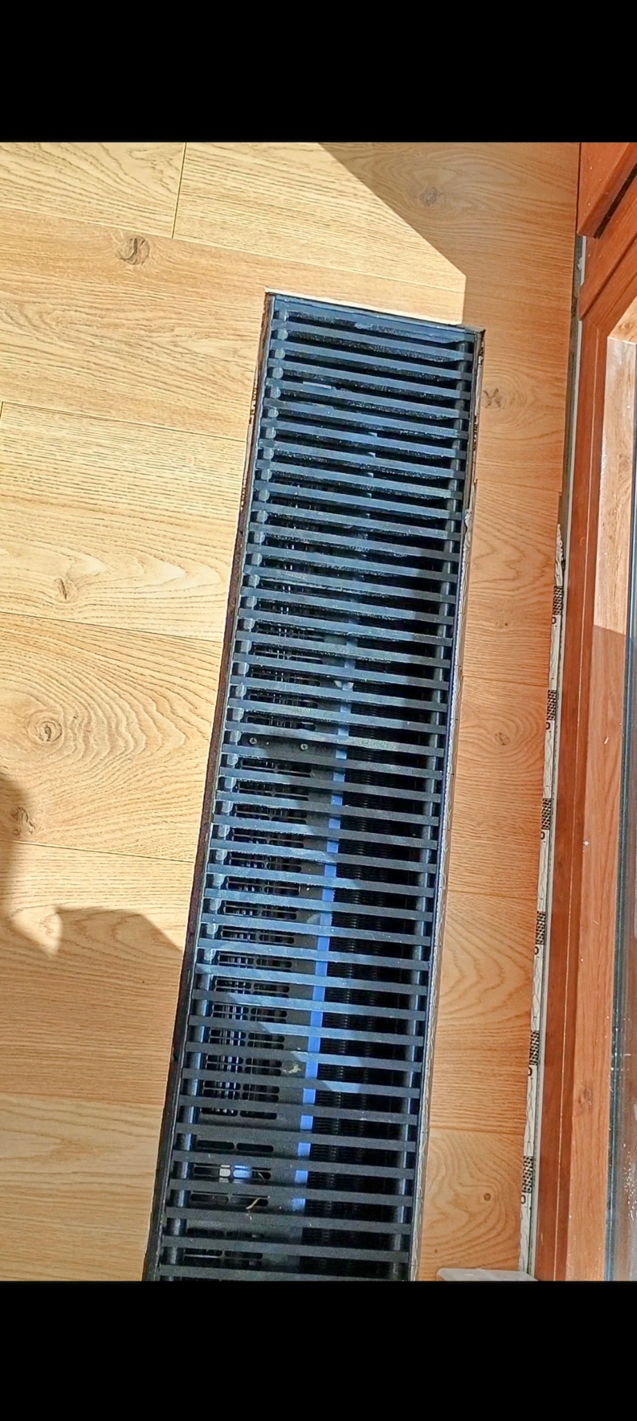 Szczegółowe ujęcie liniowego odpływu podłogowego wkomponowanego w jasne panele podłogowe, z widoczną krawędzią okna po prawej stronie.