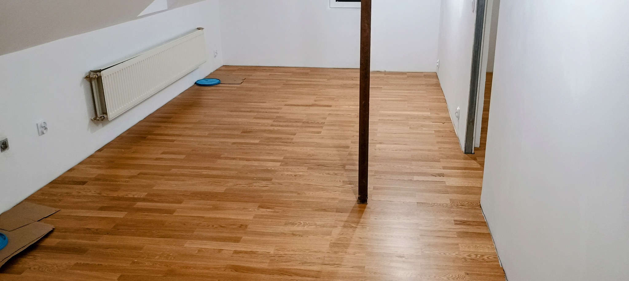 Pokój na poddaszu z nowo ułożoną podłogą z paneli imitujących drewno, widoczny grzejnik na ścianie, pionowa rura i fragment kartonu z niebieskim krążkiem.