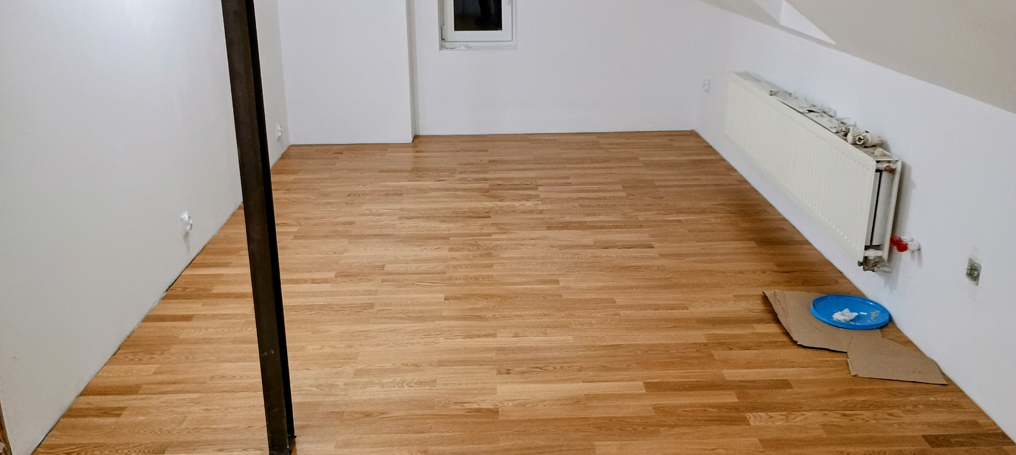 Nowa podłoga z paneli w jasnym odcieniu drewna w pokoju z białymi ścianami i grzejnikiem. Widoczne akcesoria malarskie na podłodze, sugerujące prace wykończeniowe.