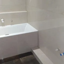 Nowoczesna łazienka z wanną obudowaną jasnymi płytkami ceramicznymi, ciemna podłoga z płytek imitujących kamień, widoczny fragment instalacji wodnej.