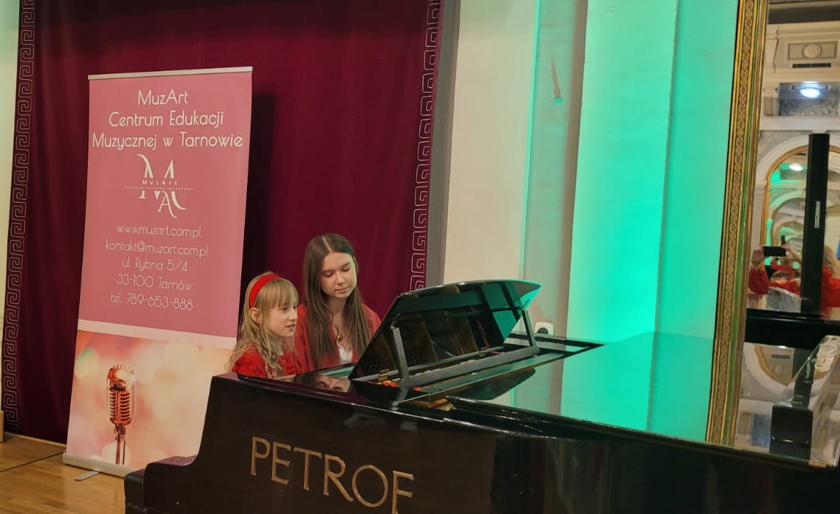 Dwie dziewczynki siedzą przy czarnym fortepianie marki Petrof, obok stoi roll-up z nazwą MuzArt Centrum Edukacji Muzycznej w Tarnowie.