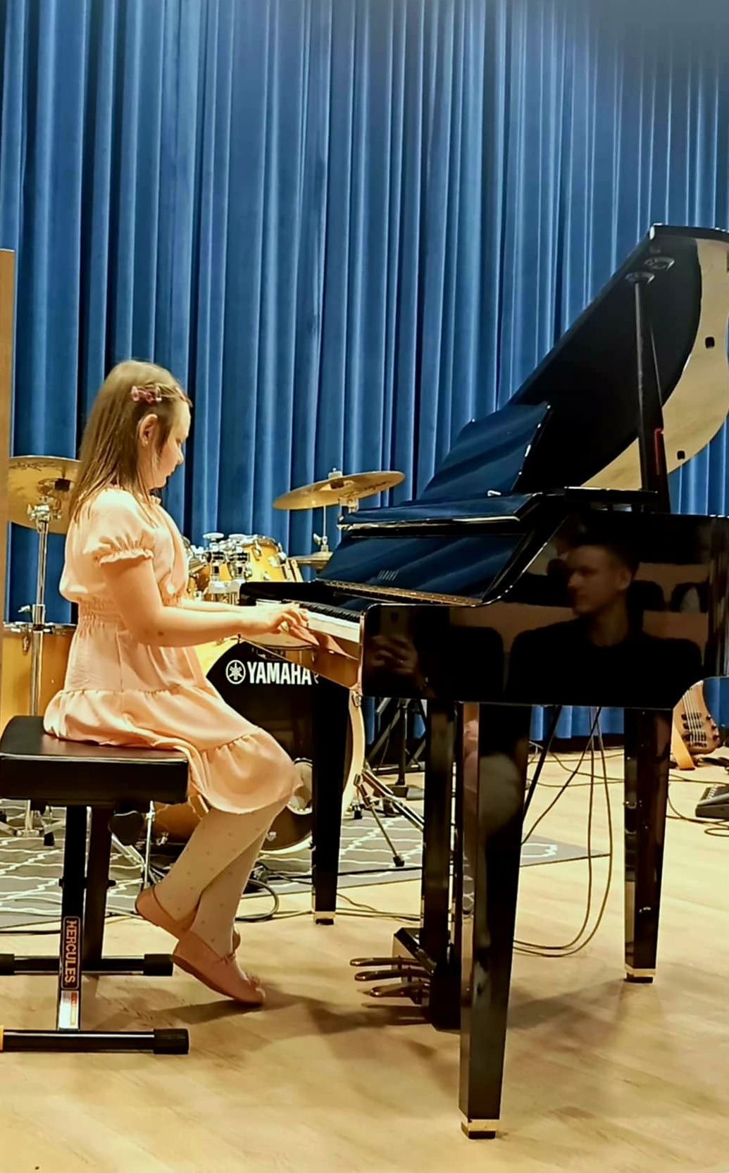 Dziewczynka w różowej sukience gra na czarnym fortepianie Yamaha w sali z błękitnymi zasłonami i perkusją w tle.