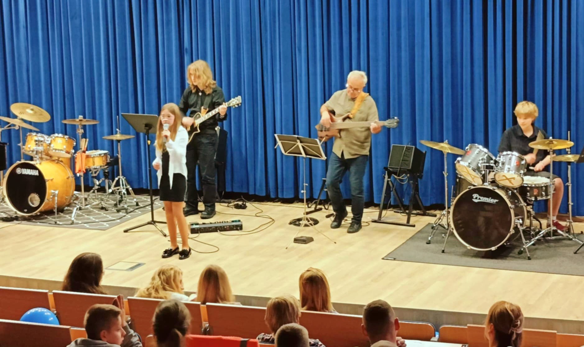 Zespół muzyczny z młodą wokalistką na scenie podczas występu. W tle błękitna kurtyna i widownia. Perkusja Yamaha i Premier.