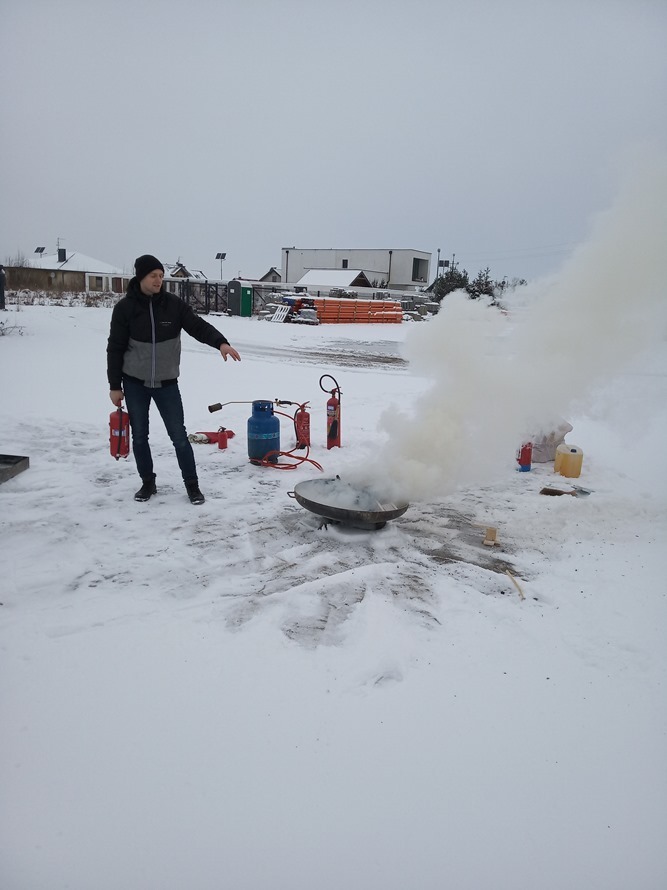 Instruktor w zimowej scenerii demonstruje gaszenie ognia w metalowej misie, używając gaśnicy i palnika, z widoczną butlą gazową i dymem.