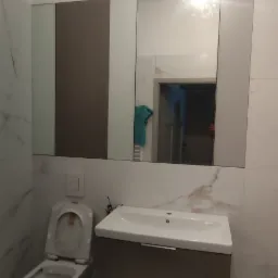 Nowoczesna łazienka z toaletą, umywalką z szafką w kolorze taupe i lustrem z elementami w kolorze taupe, widoczne kable wystające z sufitu.