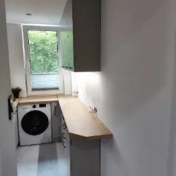 Szara zabudowa z drewnianym blatem w małej łazience z pralką; widoczne wystające kable elektryczne.