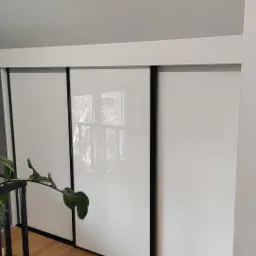 Biała szafa przesuwna z czarnymi obramowaniami, widoczna z boku, z rośliną doniczkową w kadrze, odbicie okna w drzwiach.