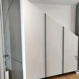 Biała szafa wnękowa z czarnymi, pionowymi akcentami, wkomponowana w nowoczesne wnętrze z szarą ścianą o geometrycznym wzorze. Widoczny fragment drewnianej podłogi i zielona roślina doniczkowa.