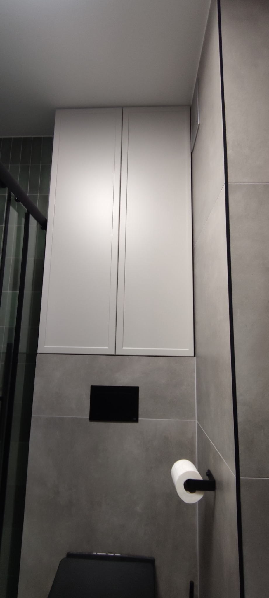 Biała szafka wisząca nad toaletą w nowoczesnej łazience z szarymi płytkami i czarnymi akcentami.