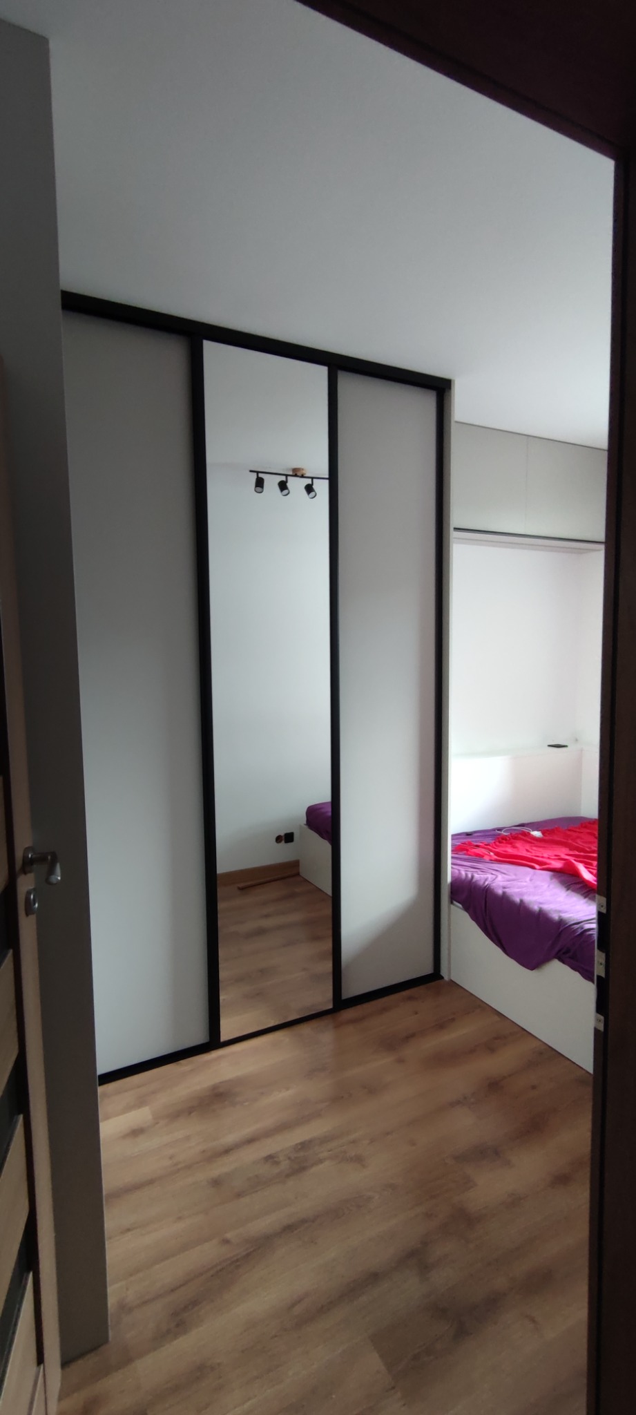 Nowoczesna szafa wnękowa z przesuwnymi drzwiami i lustrem w czarnej ramie, widoczna w sypialni z drewnianą podłogą i łóżkiem w zabudowie.