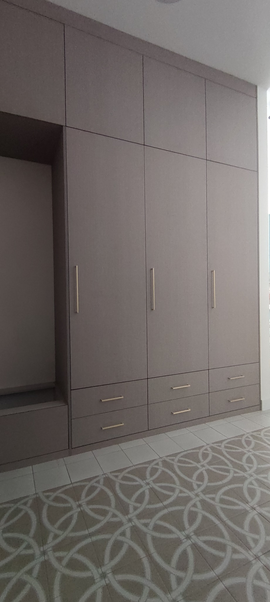 Duża, zabudowana szafa z szarymi frontami, złotymi uchwytami i otwartą wnęką, na podłodze wzorzyste płytki.