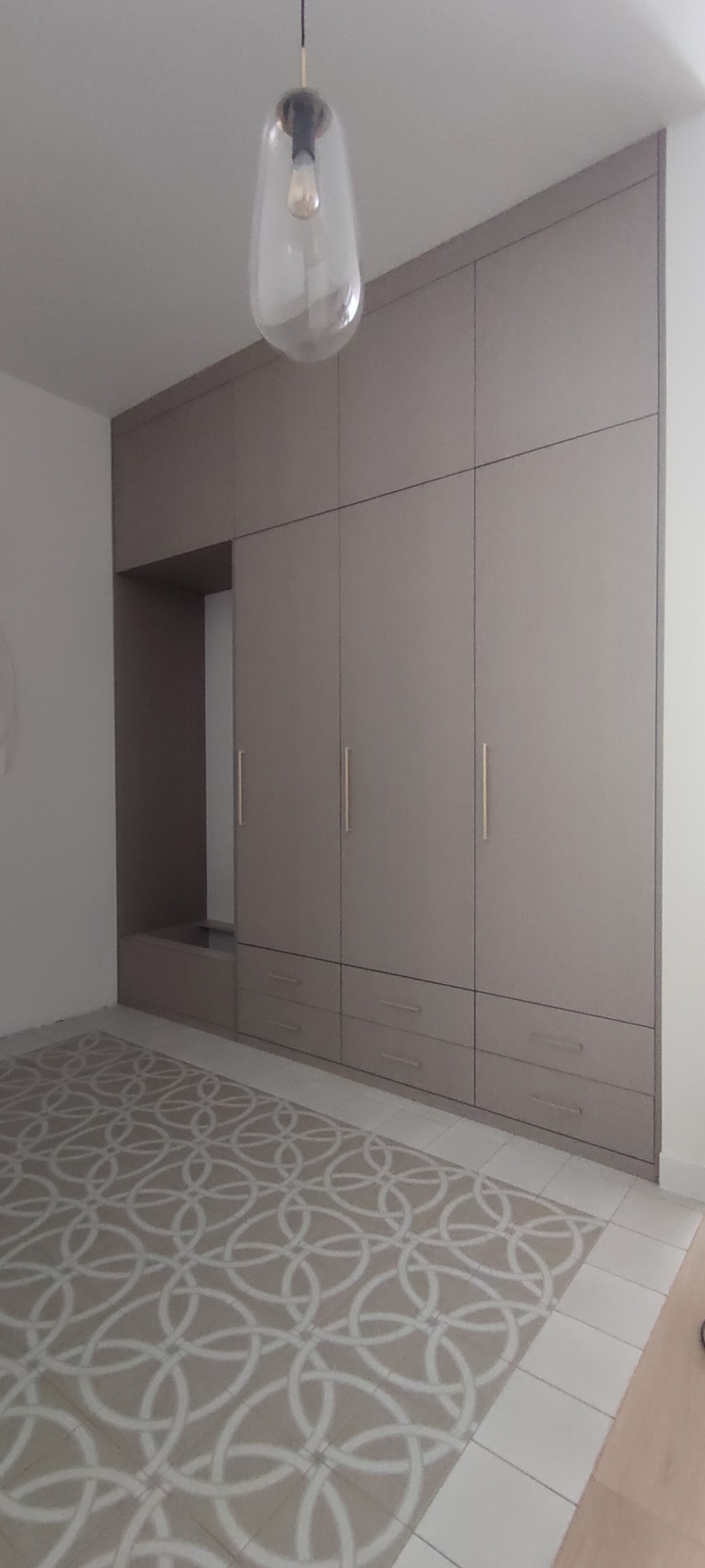 Duża, zabudowana szafa w neutralnym kolorze z otwartą wnęką, oświetlona wiszącą lampą z przezroczystego szkła, na tle wzorzystej podłogi.