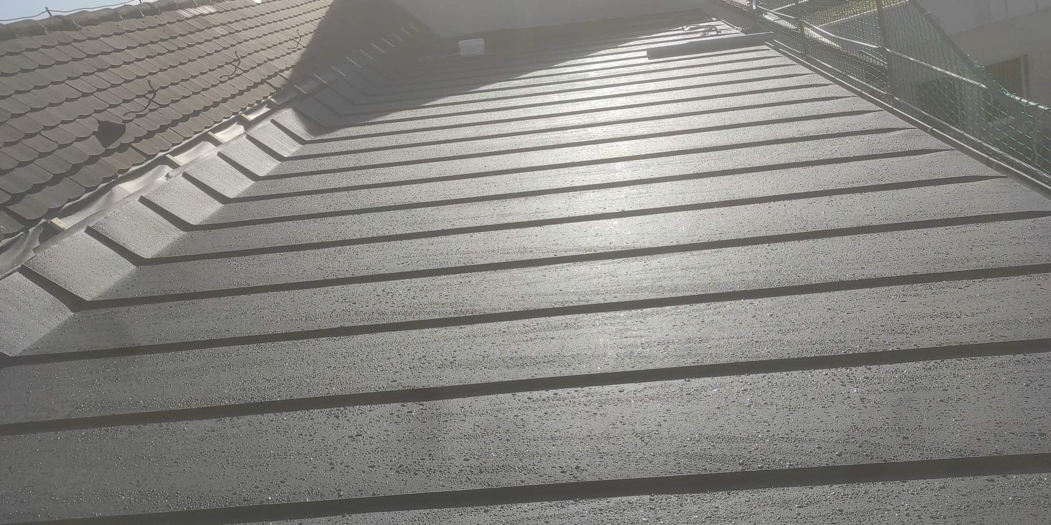 Nowy, szary dach z blachy trapezowej, pokryty drobnymi kroplami wody, sąsiadujący z fragmentem dachu pokrytego dachówką ceramiczną.