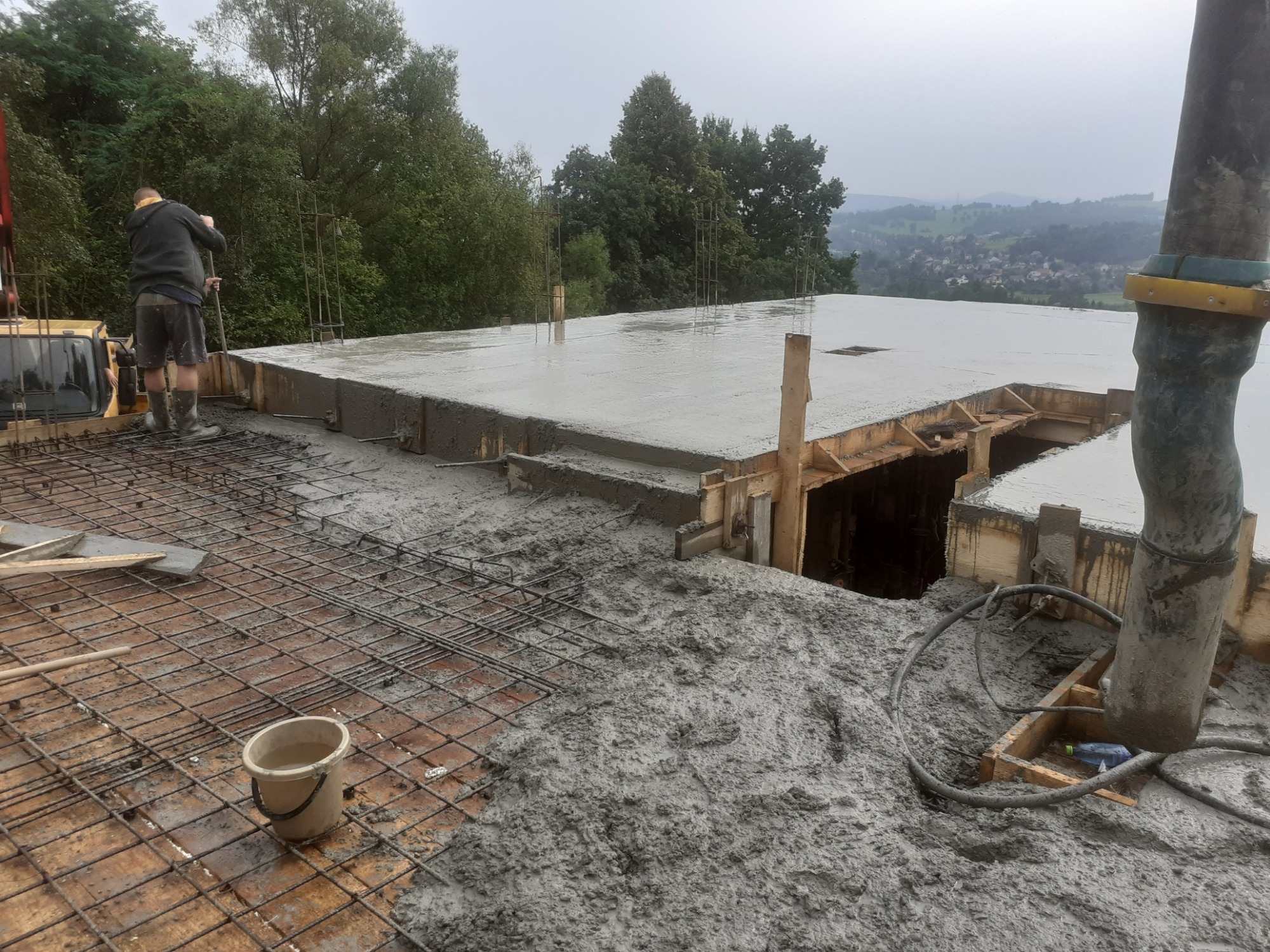 Wylewanie betonu na strop domu jednorodzinnego, widoczne zbrojenie, szalunki oraz pracownik w gumowych butach wyrównujący powierzchnię. W tle zalesione wzgórza i zabudowania.
