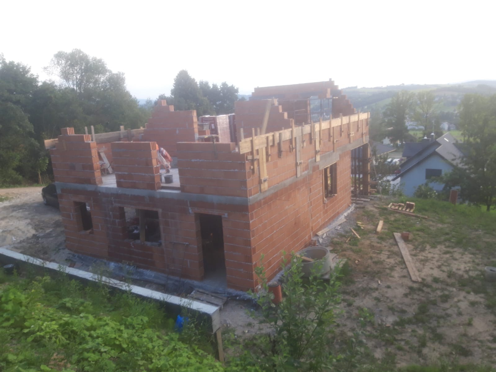 Budowa domu murowanego w stanie surowym otwartym z widoczną konstrukcją drewnianą stropu, na tle pagórkowatego krajobrazu z zabudowaniami.