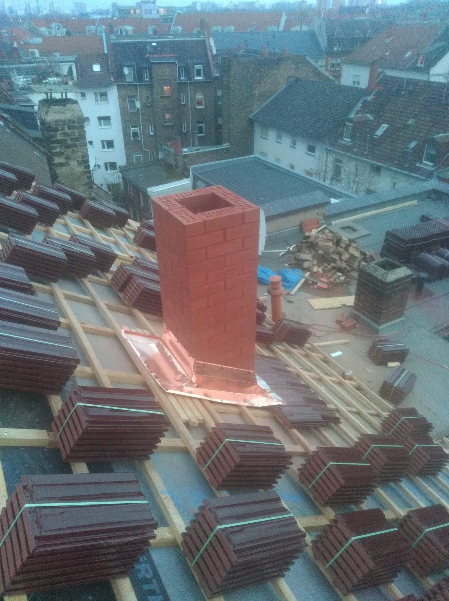 Montaż dachu: widok z góry na konstrukcję drewnianą z ułożonymi paczkami brązowej dachówki ceramicznej i nowym kominem z cegły z obróbką blacharską, w tle zabudowa miejska.