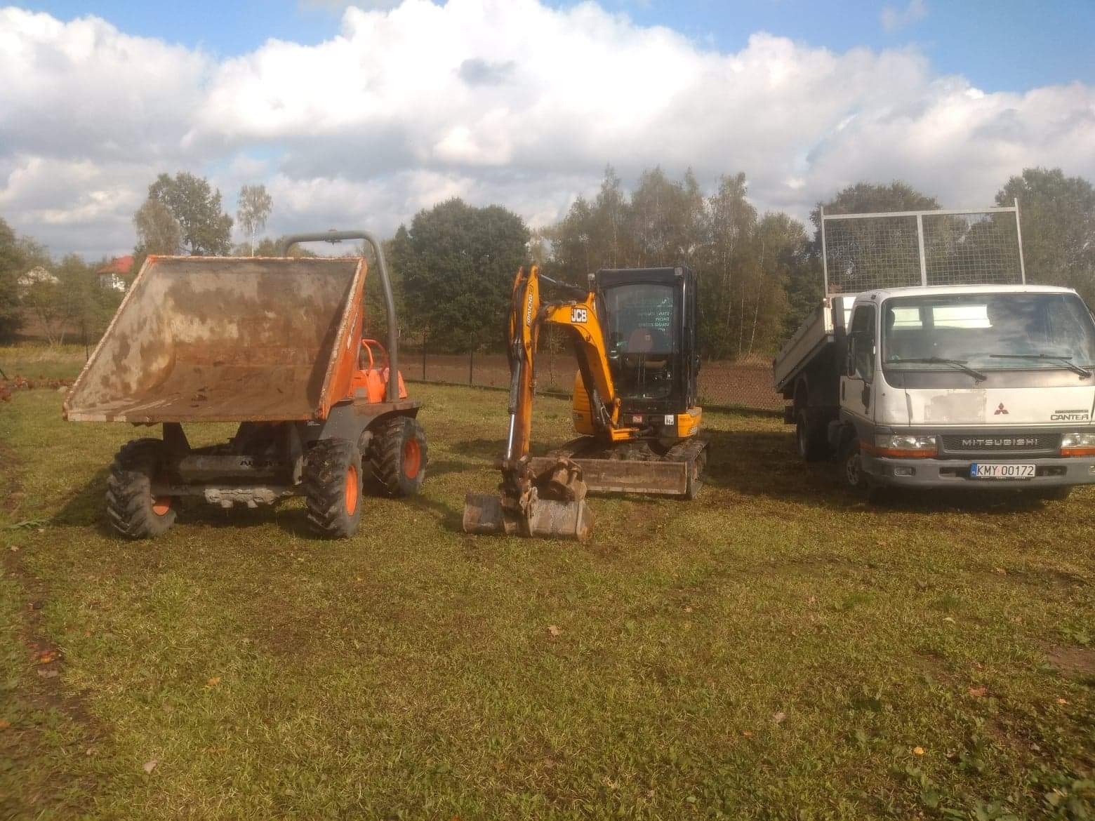 Pomarańczowa wywrotka, żółta koparka JCB i biała ciężarówka Mitsubishi Canter stoją na trawiastym terenie pod błękitnym niebem z chmurami.