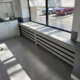 Wnętrze gabinetu kosmetycznego z szarymi płytkami podłogowymi, białymi meblami i grzejnikiem pod oknem z żaluzjami.