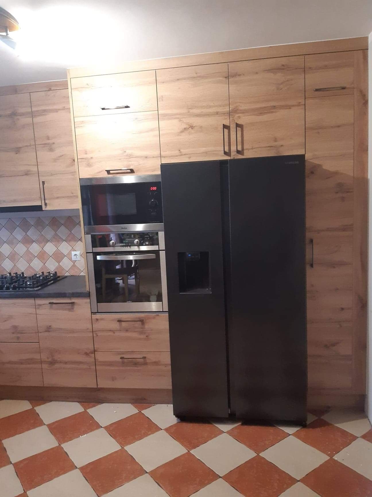 Kuchenna zabudowa z drewnopodobnymi frontami, zintegrowana z piekarnikiem, mikrofalówką i czarną lodówką side-by-side, na podłodze płytki w szachownicę.