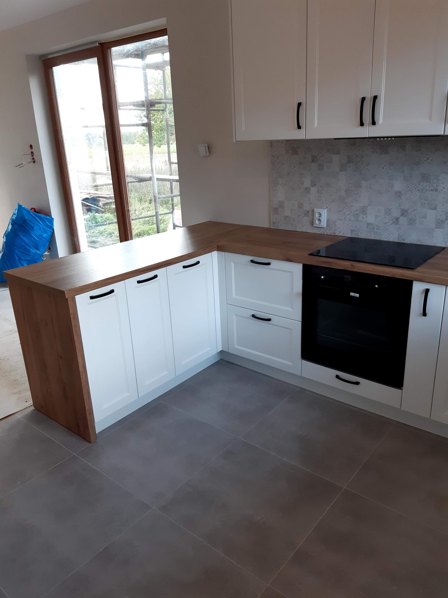 Nowoczesna kuchnia z białymi szafkami z czarnymi uchwytami i drewnianym blatem, widok na okno z rusztowaniem na zewnątrz, szare płytki podłogowe.