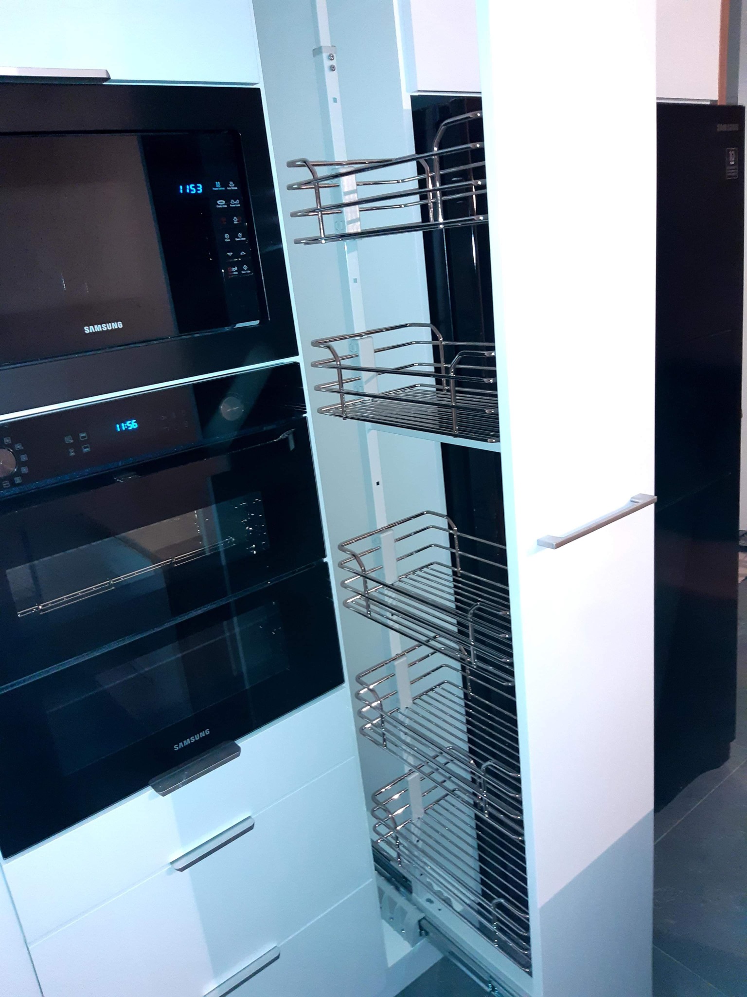 Biała zabudowa kuchenna z wysuwanym koszem cargo, obok piekarnik i mikrofalówka marki Samsung w czarnej obudowie.