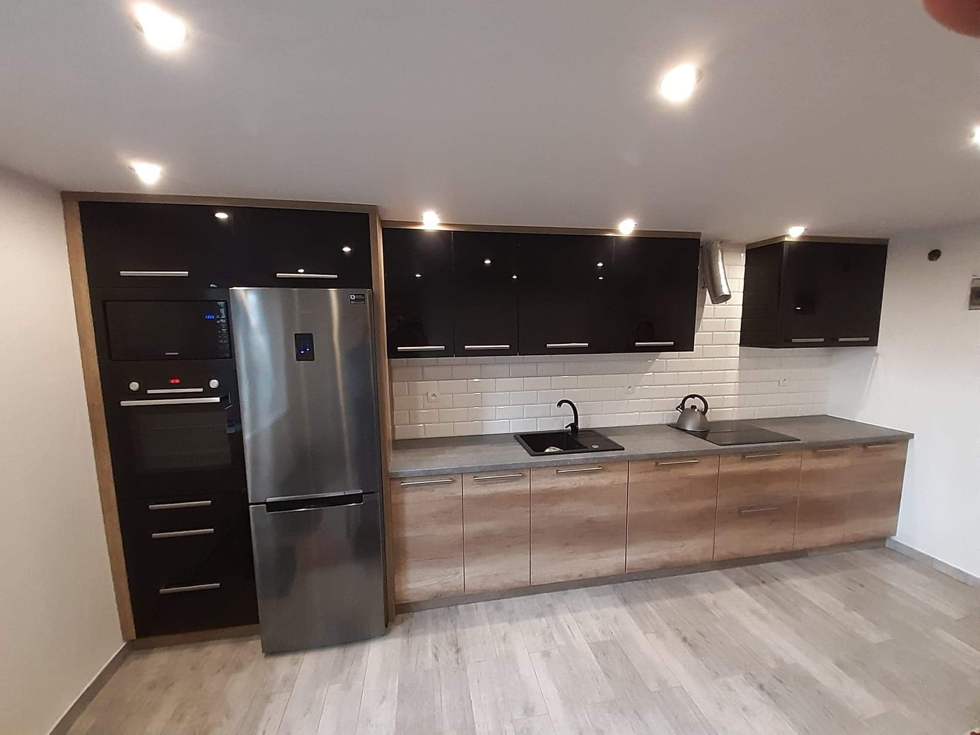 Nowoczesna kuchnia z czarnymi, błyszczącymi szafkami górnymi i drewnopodobnymi szafkami dolnymi, uzupełniona o zabudowaną lodówkę i piekarnik w czarnej obudowie.