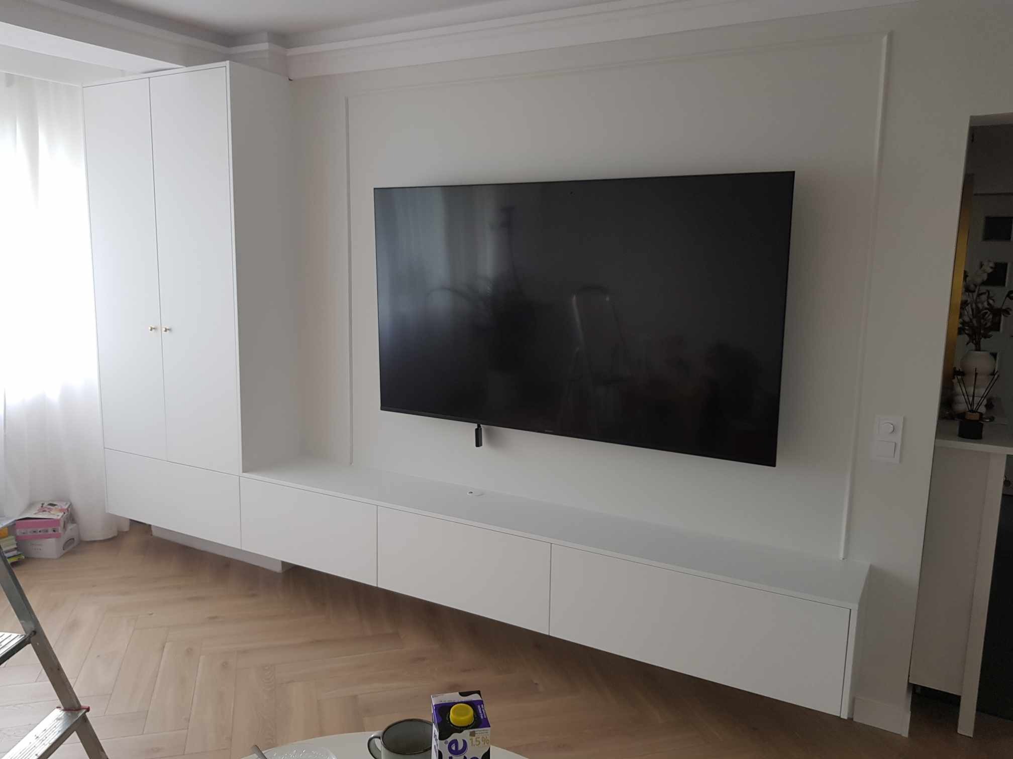 Biała zabudowa meblowa w salonie z telewizorem, obejmująca szafę i szafki RTV, na podłodze parkiet w jodełkę.