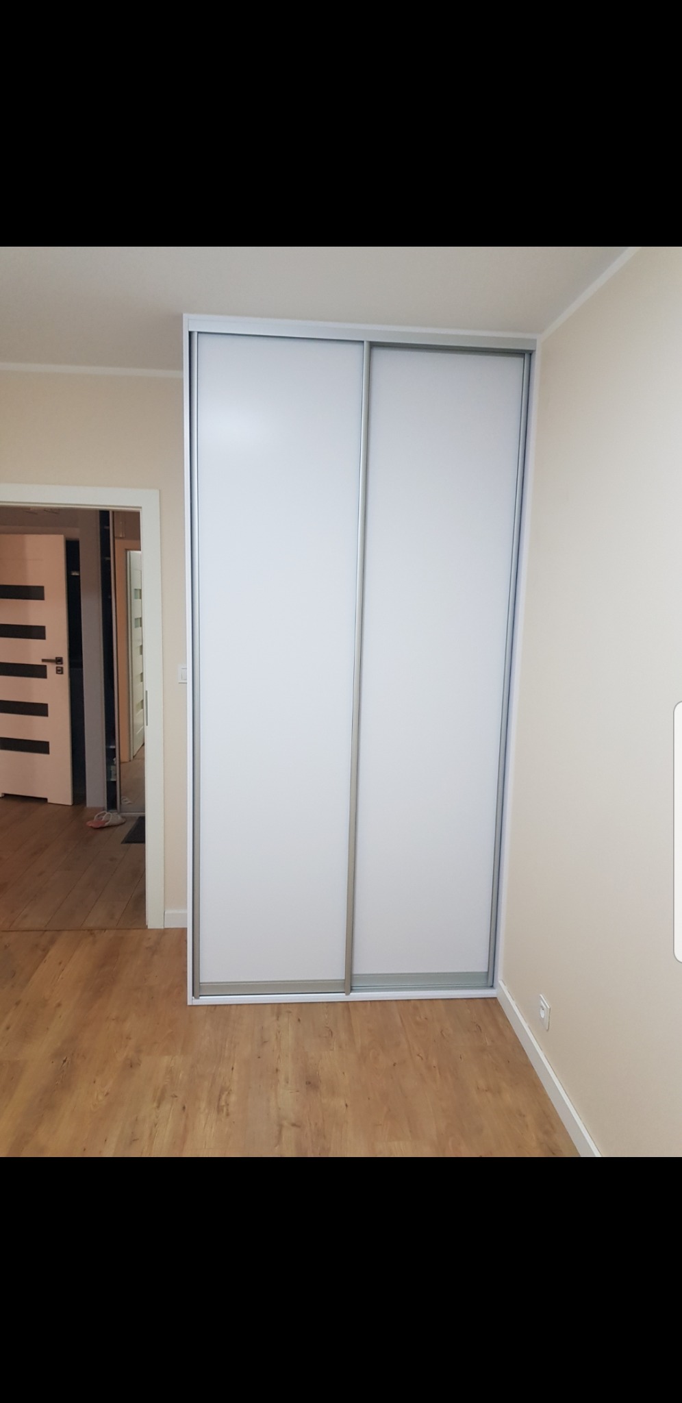 Biała szafa przesuwna z dwoma drzwiami w pokoju z panelami podłogowymi, widok na otwarte drzwi do innego pomieszczenia.