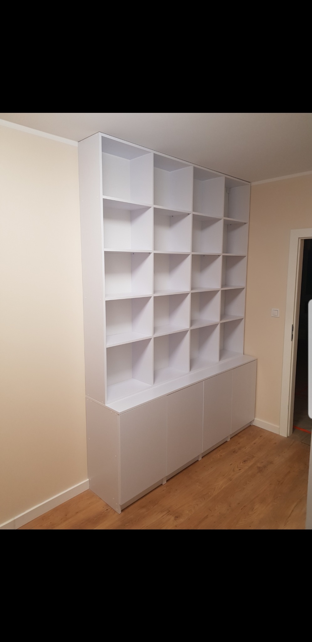 Biała zabudowa meblowa z półkami i szafkami w jasnym wnętrzu, drewnopodobna podłoga, widok na drzwi w tle.