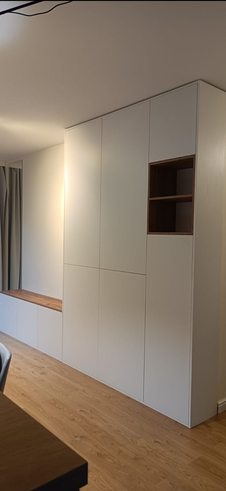 Biała zabudowa meblowa z półkami w odcieniu drewna, wkomponowana w ścianę, z drewnianym siedziskiem pod oknem i zasłonami.