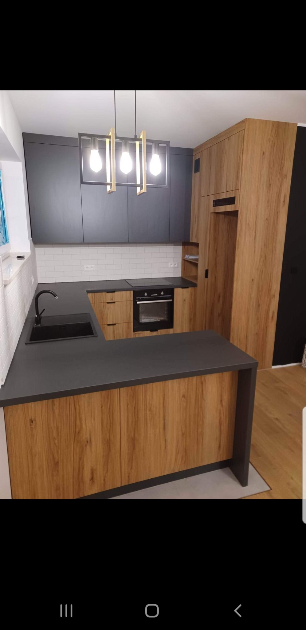 Nowoczesna kuchnia z drewnianymi frontami szafek dolnych i czarnymi szafkami górnymi, czarny blat, bateria i zlew, białe kafelki na ścianie oraz lampa wisząca z trzema żarówkami.