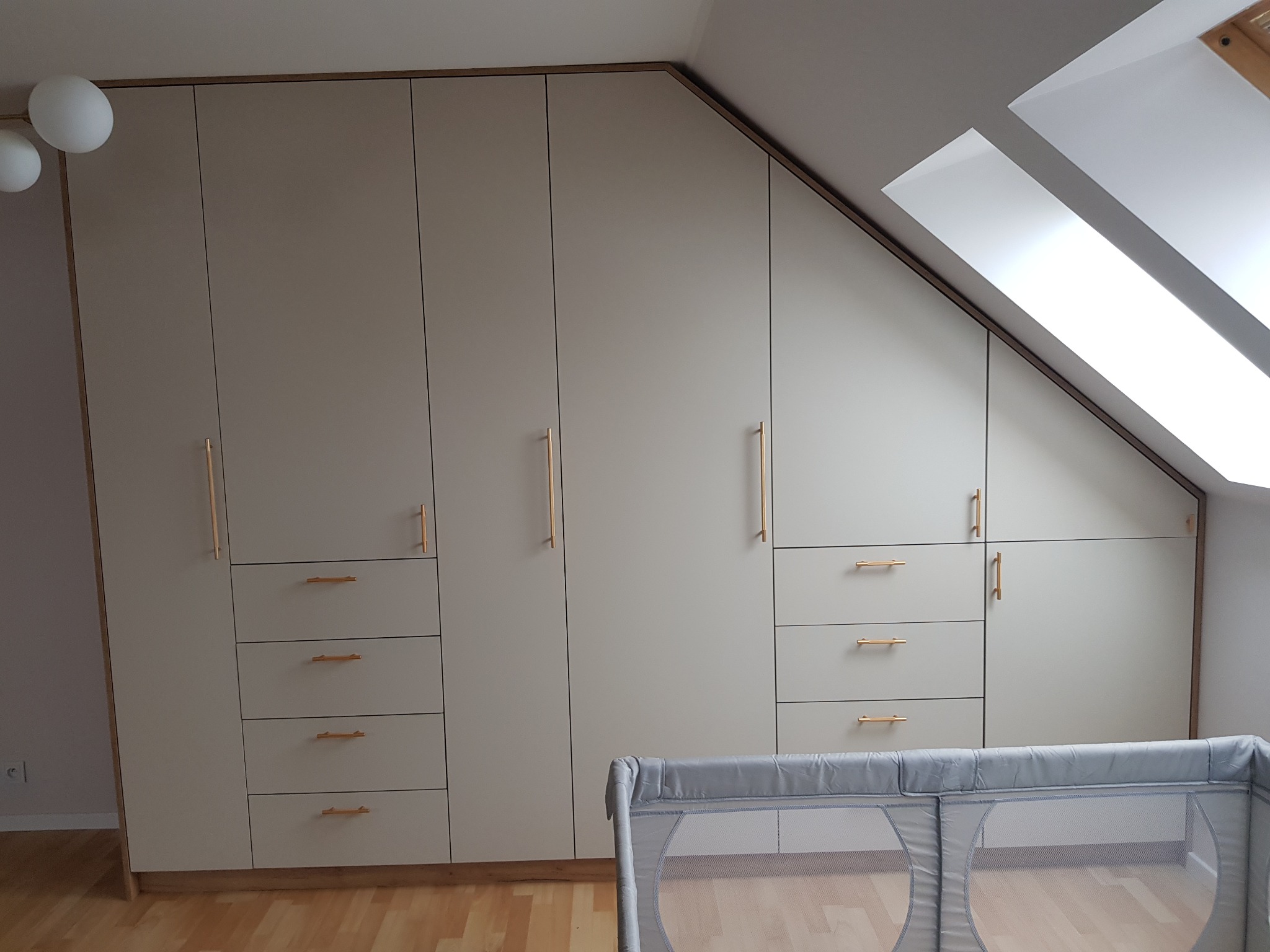 Jasnoszara zabudowa meblowa pod skosem na poddaszu, z szufladami i szafkami z podłużnymi złotymi uchwytami, widoczny fragment dziecięcego łóżeczka.