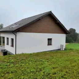 Nowoczesny, parterowy dom z białymi ścianami, brązową drewnianą górną elewacją, brązowym dachem i małymi oknami, otoczony zielonym terenem, z widoczną jednostką klimatyzacji na zewnątrz.