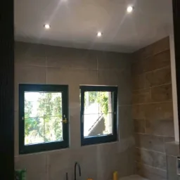 Wykończona łazienka z wanną obudowaną płytkami, dwoma oknami z widokiem na zieleń, oświetleniem sufitowym i szarą kolorystyką płytek na ścianach.