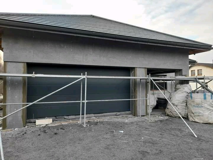 Dom w trakcie ocieplania, widoczne rusztowanie, szara elewacja w surowym stanie, brama garażowa i worki z materiałem budowlanym na ziemi.