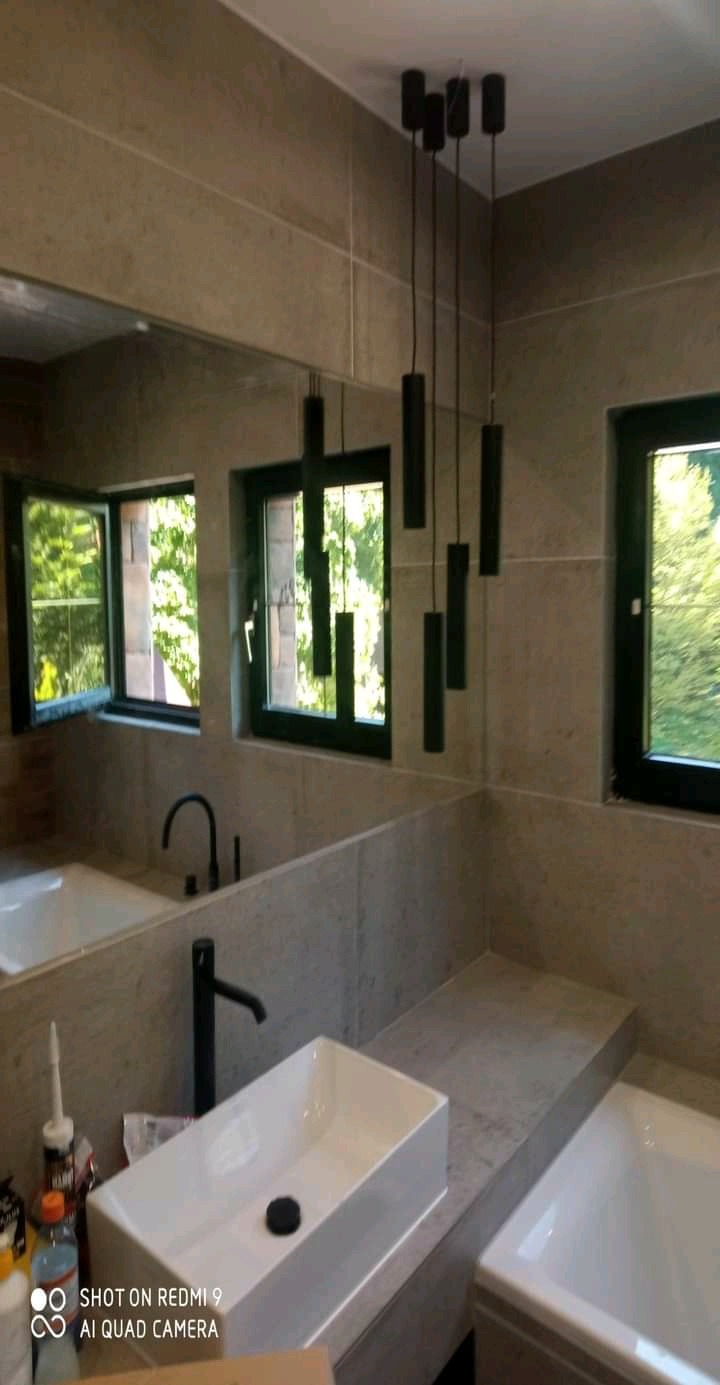 Wykończona łazienka z beżowymi płytkami, białą prostokątną umywalką, czarną armaturą i wiszącymi czarnymi lampami cylindrycznymi. Widoczne okna z zielenią na zewnątrz.