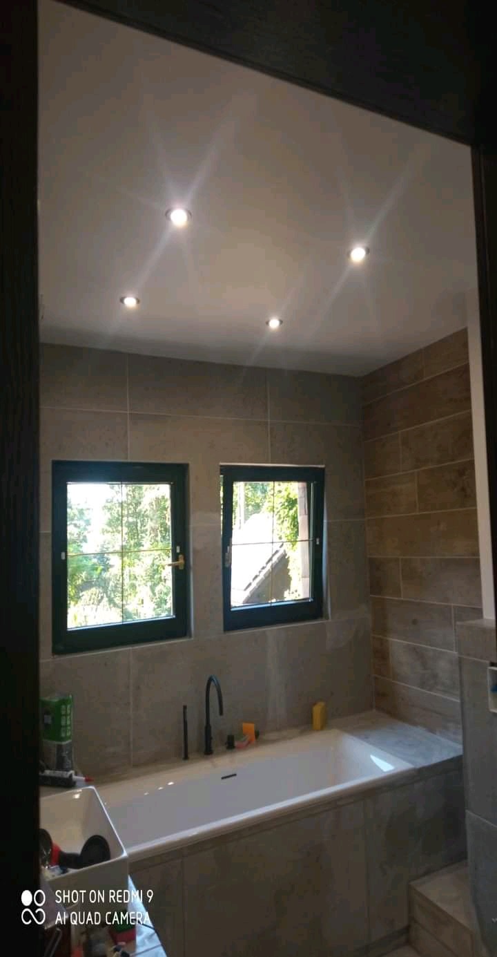 Wykończona łazienka z wanną obudowaną płytkami, dwoma oknami z widokiem na zieleń, oświetleniem sufitowym i szarą kolorystyką płytek na ścianach.