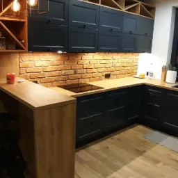 Nowoczesna kuchnia z ciemnymi szafkami, ceglaną ścianą i drewnianym blatem oraz podłogą, oświetlona wiszącymi lampami i punktowymi światłami.