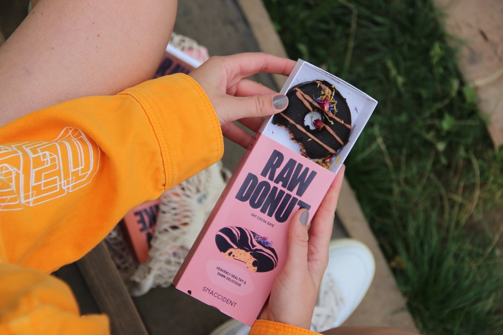 Kobieta w pomarańczowej bluzie prezentuje pudełko z napisem 'RAW DONUT' i wegańskiego donuta z czekoladą i kwiatami, marki The Snaccidents.