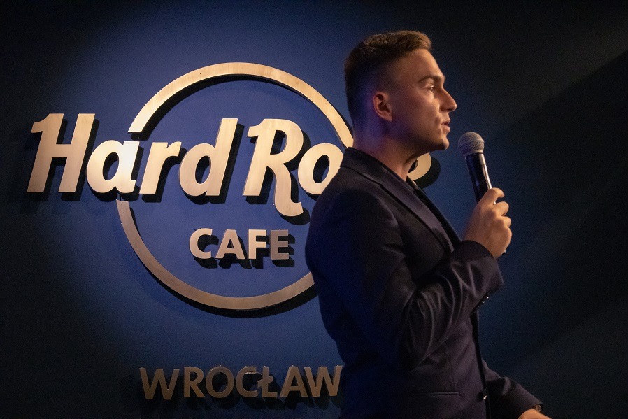 Prelegent z firmy Aries Agency z Gdańska, Oskar Król, przemawia na scenie CRASH Mondays we Wrocławiu, na tle logo Hard Rock Cafe.