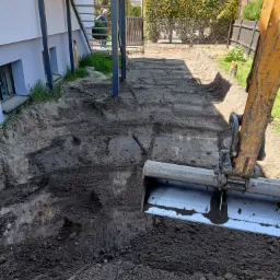 Żółta koparka z łyżką w trakcie wyrównywania terenu przy budynku z metalowymi schodami zewnętrznymi, widoczne świeże ślady opon i wykopana ziemia.