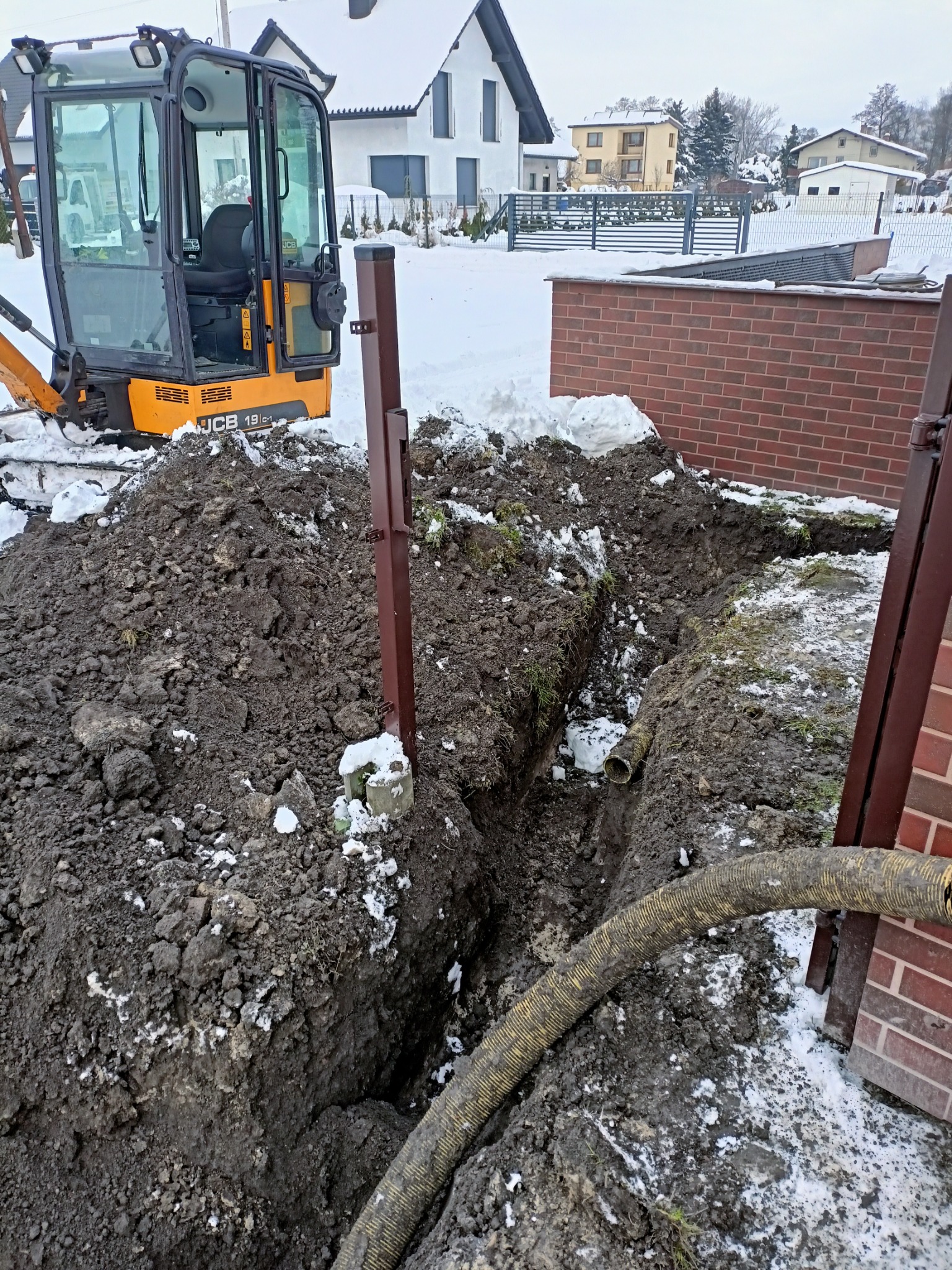 Wąski wykop w ziemi z widoczną rurą drenażową, obok stoi mini-koparka JCB 19C-1, w tle dom jednorodzinny w zimowej scenerii.